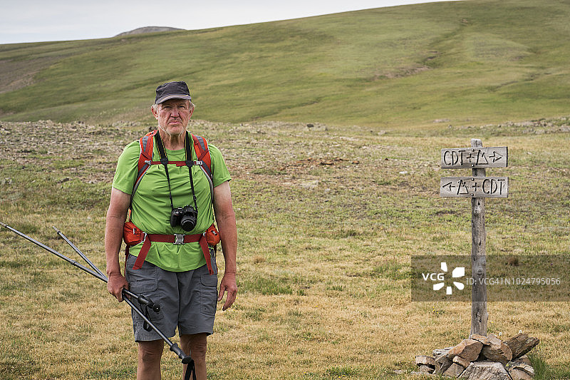 年长的男人穿着远足短裤和T恤，手持登山杖，脖子上挂着相机，站在科罗拉多步道和大陆分水岭步道的标志旁，在圣胡安山脉，韦米努切荒野，落基山脉徒步旅行图片素材