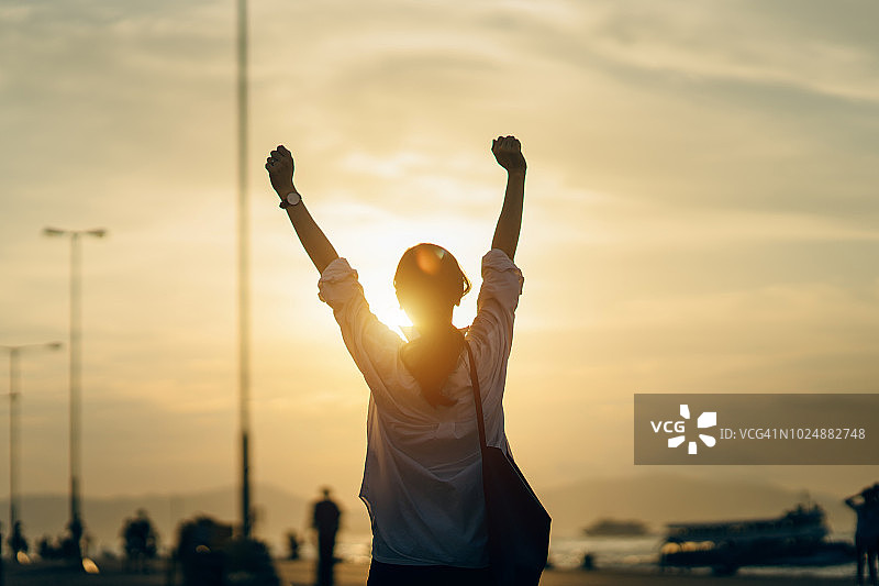年轻女子在码头边举起双手，享受美丽的日落和阳光图片素材