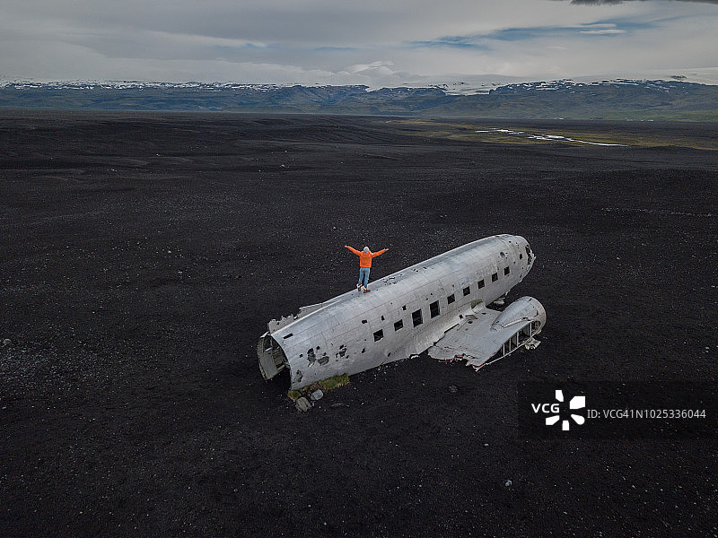年轻女子站在坠毁在黑沙滩上的飞机上，张开双臂环顾四周无人机航拍图片素材