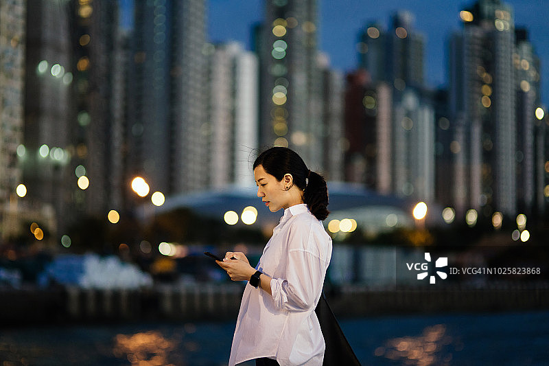 夜晚，繁华都市中在城市高楼前使用智能手机的忙碌女性图片素材