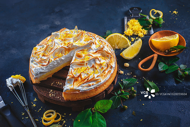 深色背景下的蛋白柠檬挞，配有树叶、柠檬皮、刮刀和搅拌器。缺少一块的馅饼。自制甜点概念图片素材