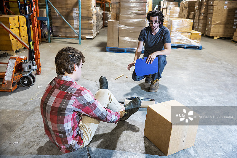 仓库工人被碎片绊倒后摔倒，主管正在帮助他，他痛苦地抓住脚踝。图片素材