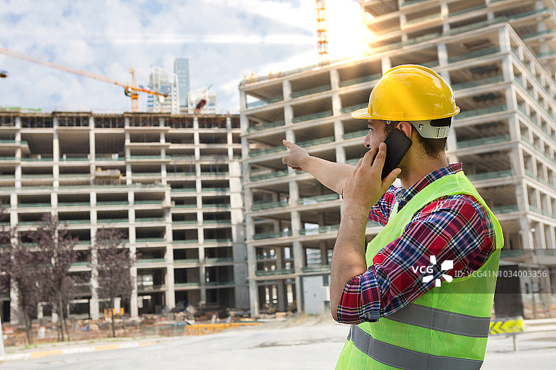 年轻工程师在建筑工地与人通电话，背景是起重机图片素材