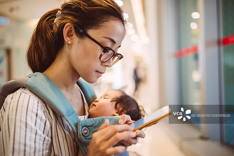 年轻妈妈用智能手机，同时用婴儿背带抱着睡着的宝宝图片素材