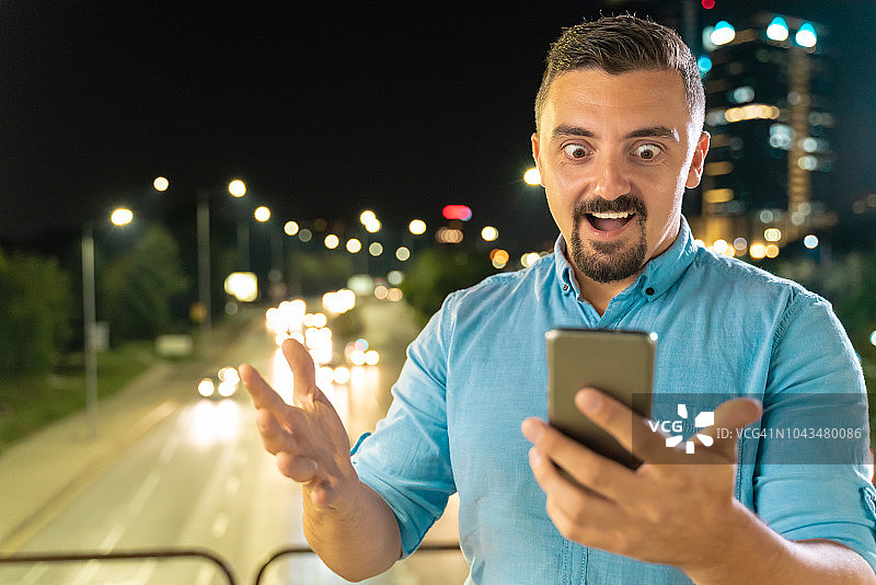 夜晚，男子在户外使用手机，面部表情惊讶，背景是城市交通图片素材