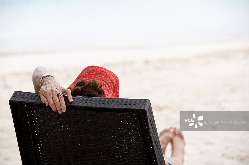 在热带岛屿度假，躺在日光躺椅上的年长女士。从后方和侧面看。一只手抬起扶住躺椅的靠背。焦点在前景的帽子和头部。框架顶部有沙子和水的复制空间图片素材