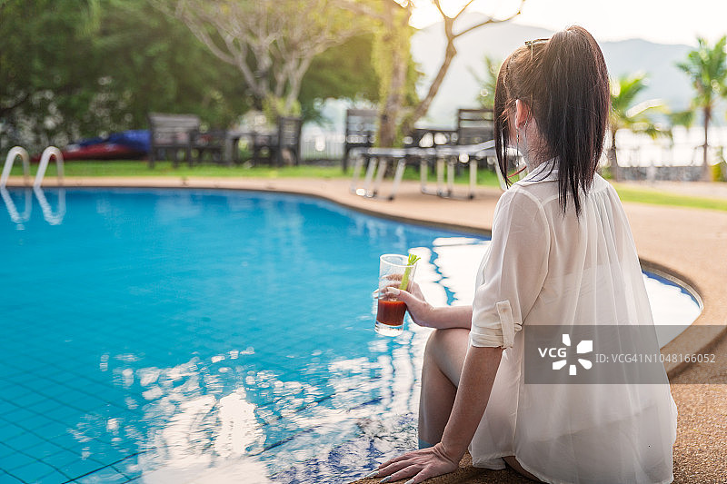 在泳池边拿着饮料，放松地看着海滩的快乐女人图片素材