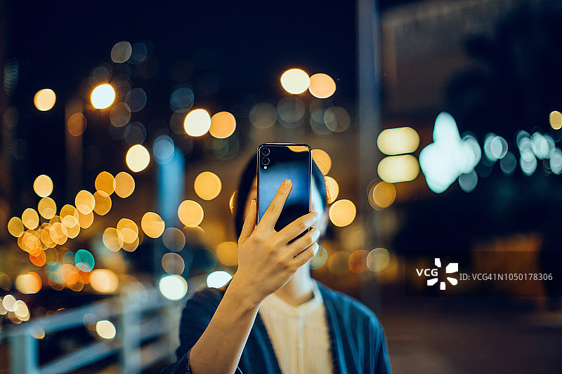 年轻亚洲女人在城市街道用智能手机拍照，背景是灯火通明的城市街道和交通图片素材