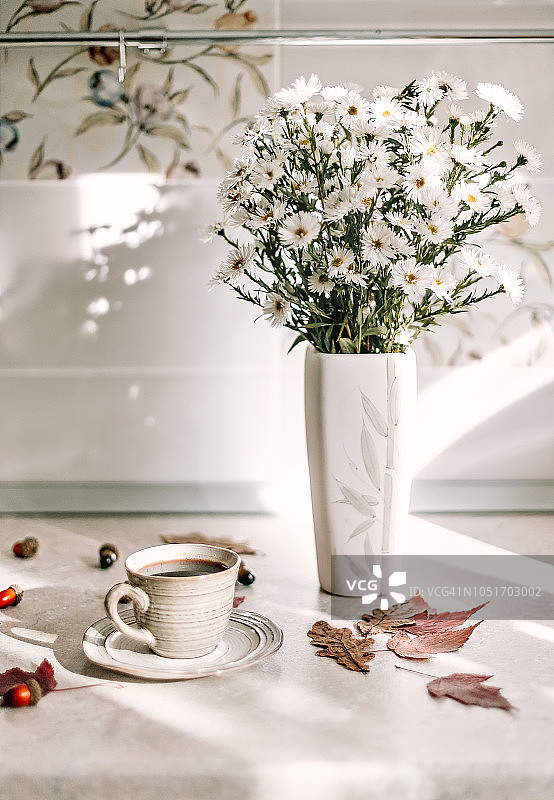 阳光穿过窗户照进厨房，白色紫苑花瓶和咖啡杯摆放在大理石桌上图片素材