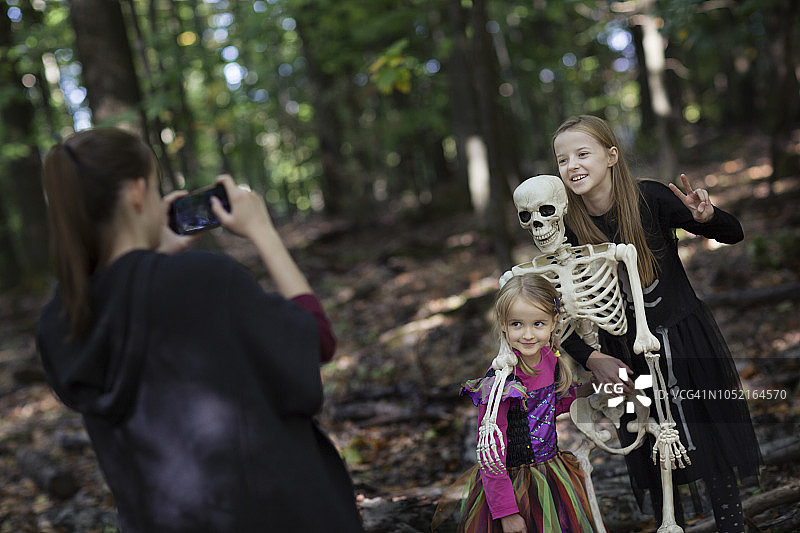 青少年用手机拍摄两个穿着万圣节服装、与森林里的假骷髅合影的孩子图片素材