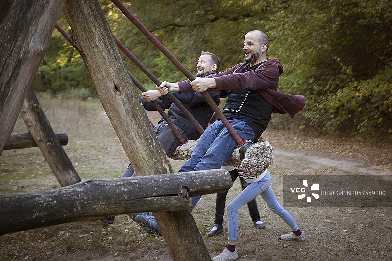 两个男人在森林游乐场玩绳索秋千，孩子们在后面推图片素材