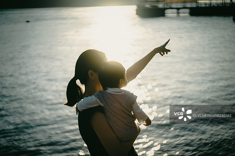 日落时分，母亲抱着女婴在海滩上指向天空图片素材