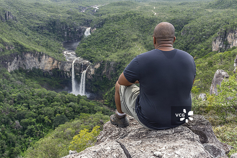 中年徒步旅行者坐在岩石边，欣赏着美丽的塞拉多景观，绿色森林中有瀑布和河流图片素材