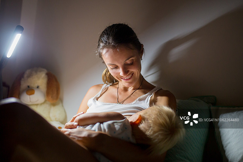 年轻美丽的母亲在昏暗灯光下母乳喂养新生儿。图片素材