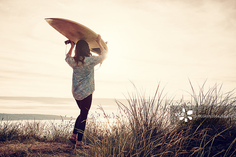 女人日落时分头顶冲浪板，海滩沙丘图片素材