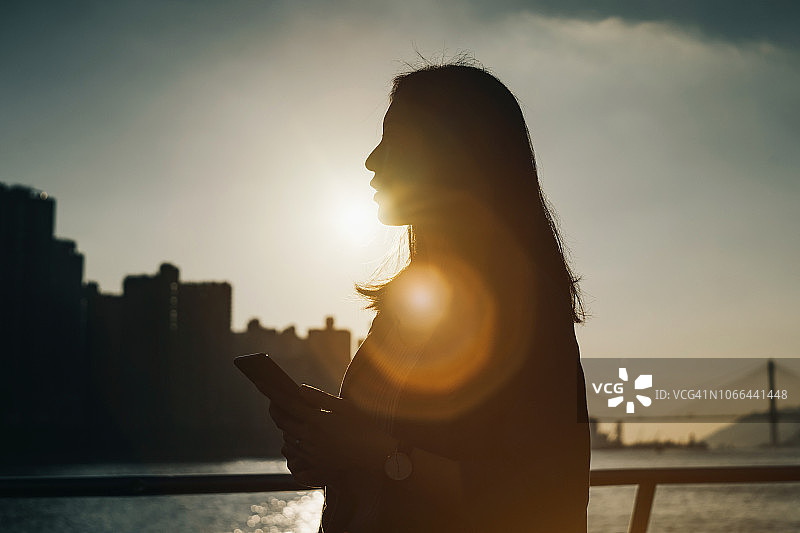 在金色日落下，于城市天际线和海港前使用手机的女子剪影图片素材