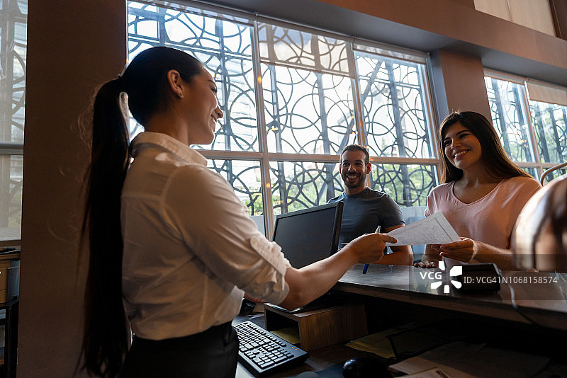 美丽女人和伴侣在酒店办理入住手续，向接待员递交表格，笑容满面图片素材