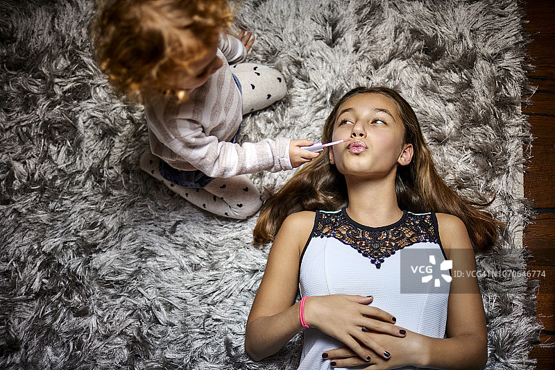 幼儿女孩为躺在长毛绒地毯上的姐姐涂口红图片素材
