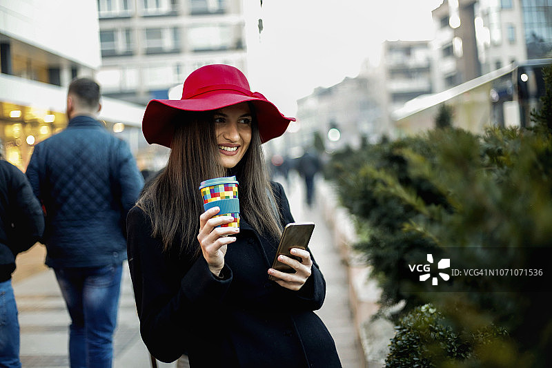 年轻女人戴着红帽子在街上喝咖啡和发短信图片素材