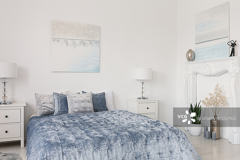 白色卧室中，蓝色床和床头柜上的灯之间的枕头，配有海报图片素材