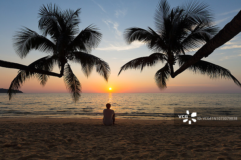 阁骨岛：男子在海滩日落时分放松地坐在倾斜向大海的两棵椰子树下图片素材