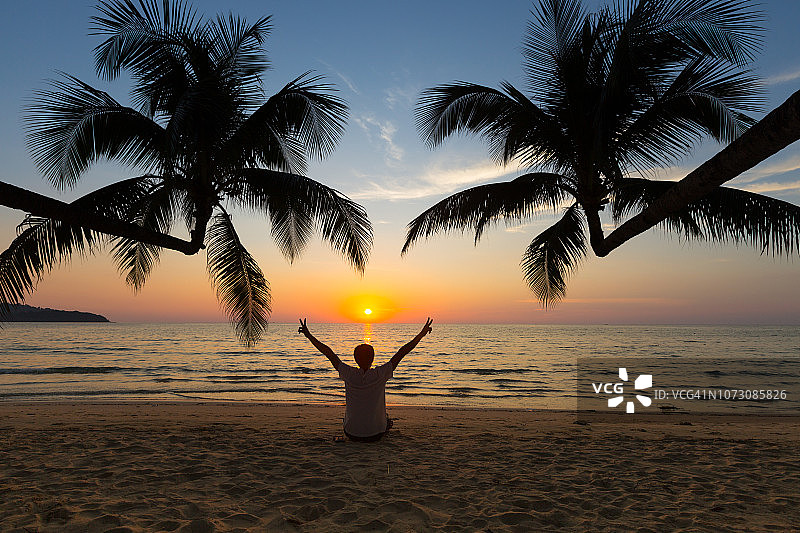 阁骨岛：男子在海滩日落时分放松地坐在倾斜向大海的两棵椰子树下图片素材