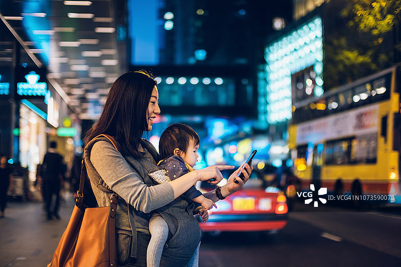 年轻的亚洲母亲用手机App在市中心街道安排出租车，背景是模糊的交通场景图片素材