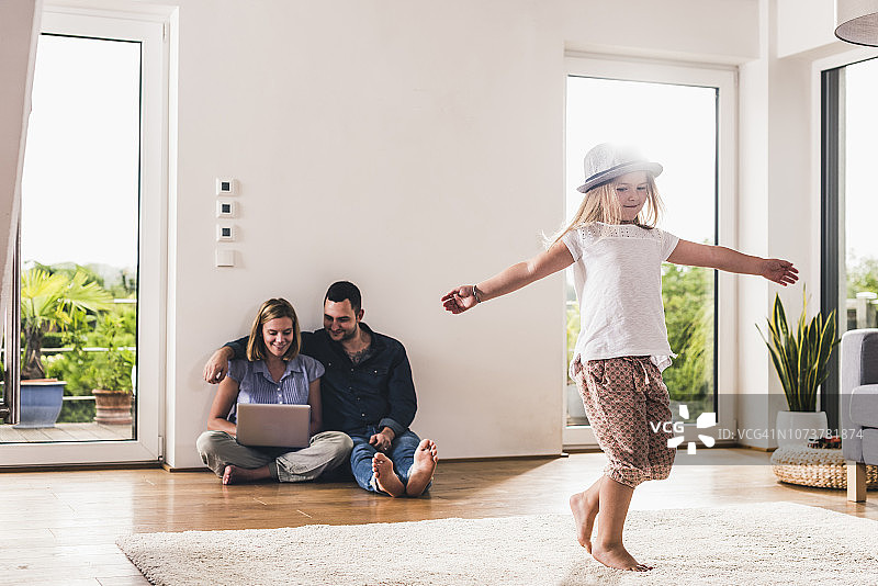 小女孩戴着帽子在家玩耍，父母在背景中使用笔记本电脑图片素材