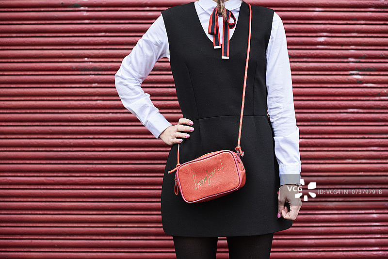 提着红色手提包、穿着黑色连衣裙的时尚女人站在红色卷帘门前图片素材