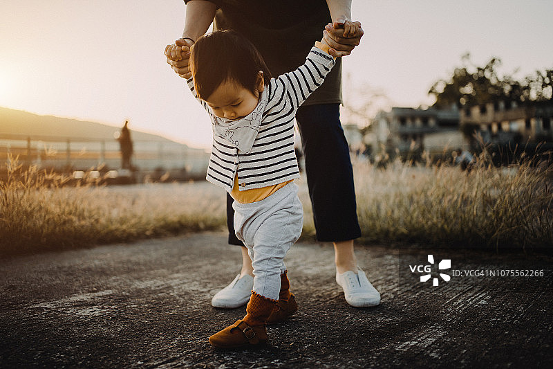 日落时分，母亲牵着女儿的手，帮助她在公园里学习走路图片素材
