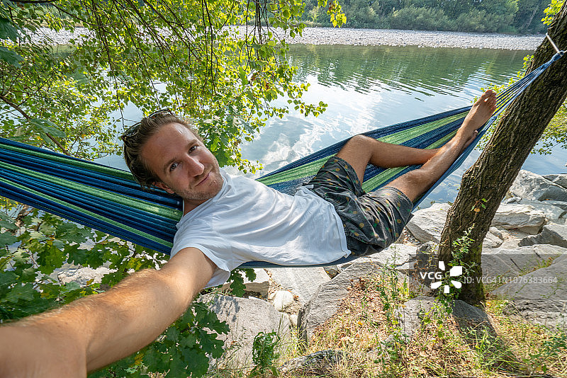 在夏天，快乐的男人在河边吊床上自拍图片素材