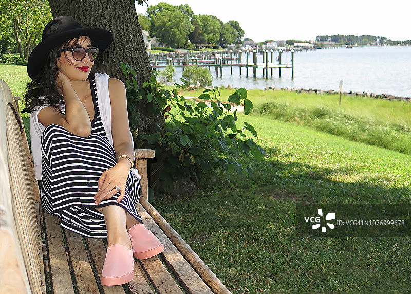 年轻女人坐在海边树下的长椅上放松和做白日梦图片素材