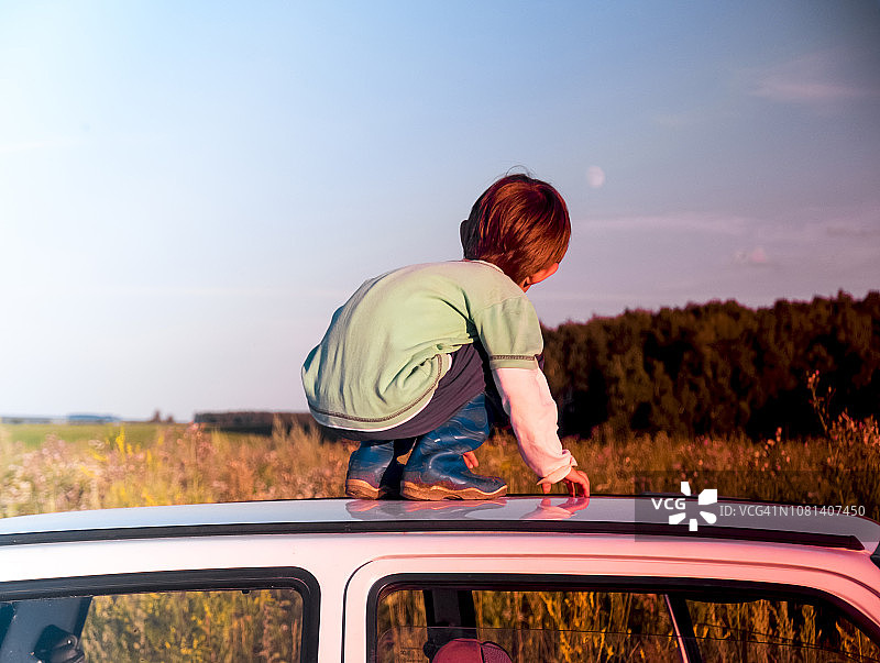 日落时分，男孩蹲在车顶上，以天空为背景的全身像图片素材