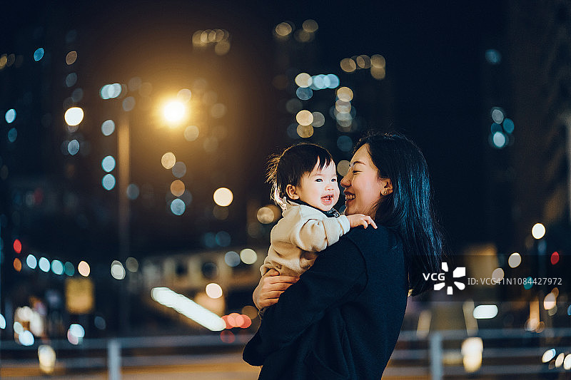 年轻的亚洲母亲在夜晚的城市里拥抱可爱的女婴，幸福地笑着图片素材