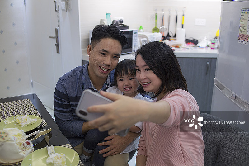 快乐的现代亚洲家庭在女儿生日庆祝活动中自拍。在香港的一家三口在公寓厨房庆祝女儿的生日并进行自拍图片素材