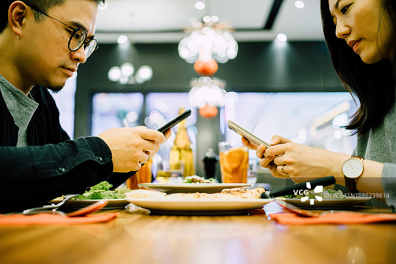 在餐厅用餐时使用智能手机且互不理睬的年轻夫妇图片素材