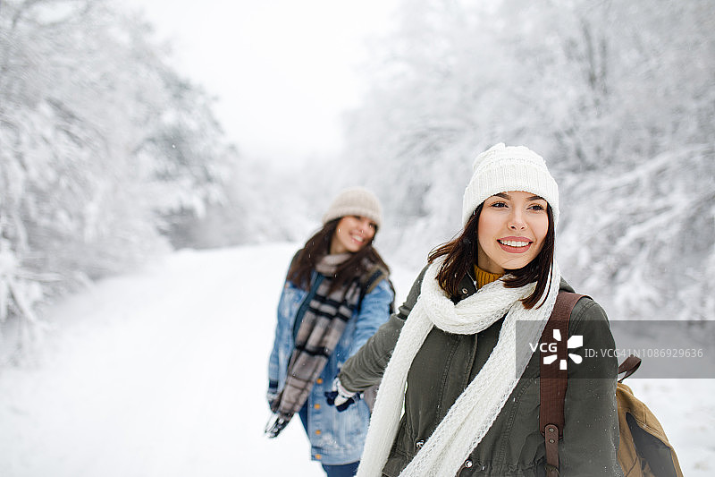 两个微笑的女人冬天在公园里散步。带着背包的朋友穿过森林。戴着针织帽和冬季夹克的女孩在白雪皑皑的森林背景下图片素材