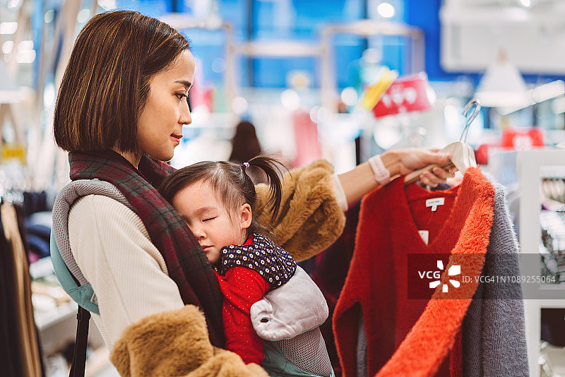 年轻漂亮的女士在时尚精品店购物时，用婴儿背带抱着睡着的婴儿，充满喜悦图片素材