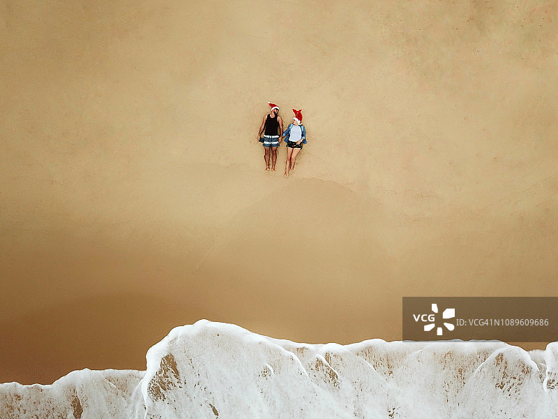 一对戴着圣诞帽的年轻情侣坐在海滩上的航拍照片图片素材