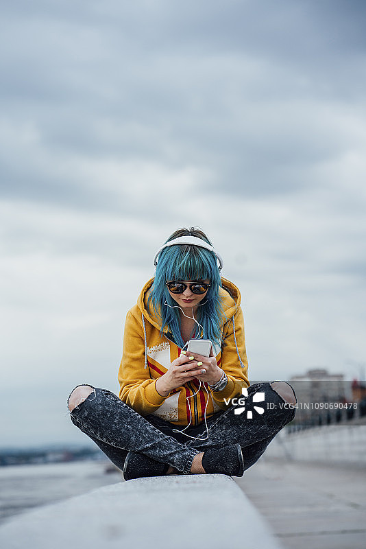 染着蓝色头发，坐在墙上用耳机和智能手机听音乐的年轻女子图片素材