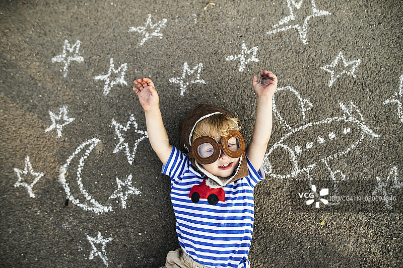戴飞行员帽和护目镜的微笑幼儿肖像，躺在绘有飞机、月亮和星星的沥青上图片素材