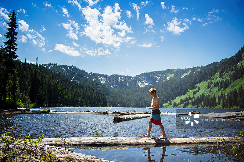 森林里，蓝天下，赤膊男孩走在湖中原木上的侧视图图片素材