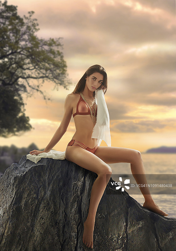穿着比基尼坐在日落岩石上的漂亮棕发女人图片素材