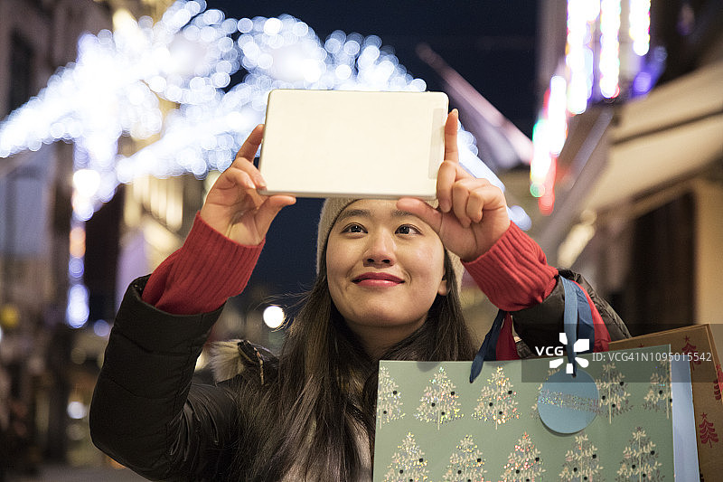 亚洲女性用平板电脑拍摄城市街道上的圣诞灯饰图片素材