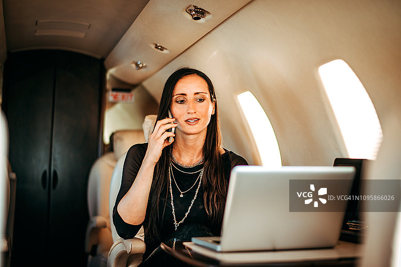 乘坐私人飞机，一边用笔记本电脑工作一边用智能手机交谈的美丽富有女人图片素材