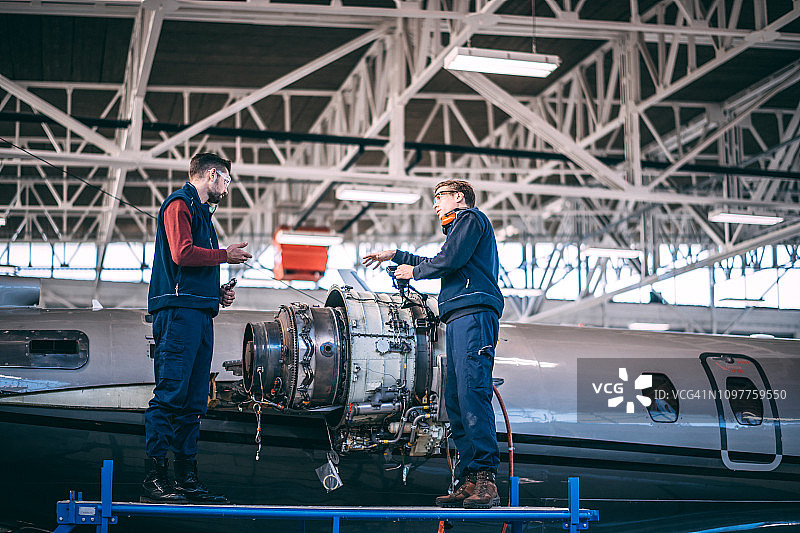 两名飞机机械师站在高架工作平台上，在维修机库中修理一架小型飞机的部分拆卸的喷气发动机时，正在交谈图片素材