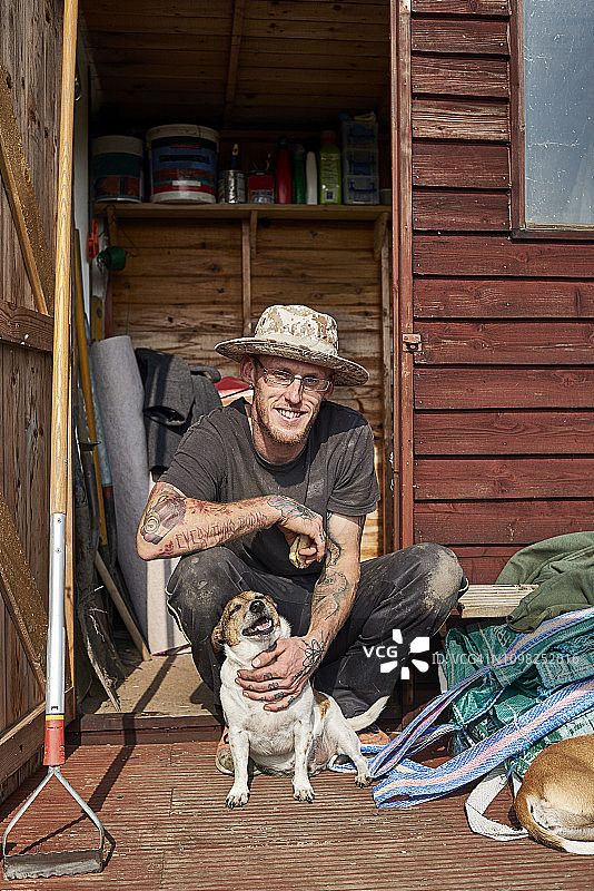 一位30多岁的英国男子在他的棚子前抚摸着他的狗的肚子图片素材