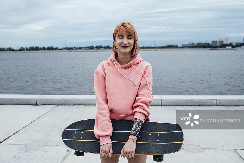 微笑的年轻女子拿着雕刻滑板站在河边图片素材