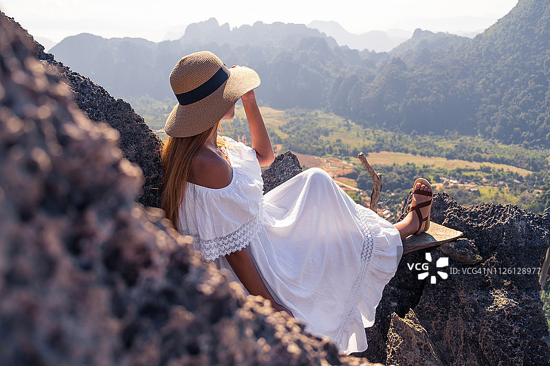 穿着白色连衣裙的女人坐在长凳上看山景，晴天图片素材