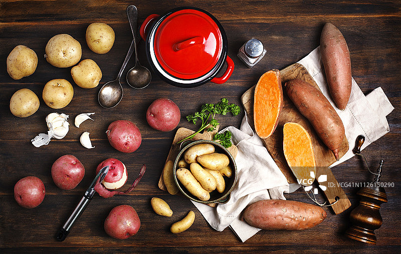 木制背景上的各种生的未煮过的有机土豆：红色、白色、甘薯和手指土豆，以及烹饪用具图片素材
